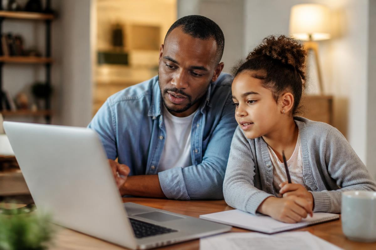 A father and his young daughter sitting at a home table, looking at a laptop and paperwork together while completing a child SSI application